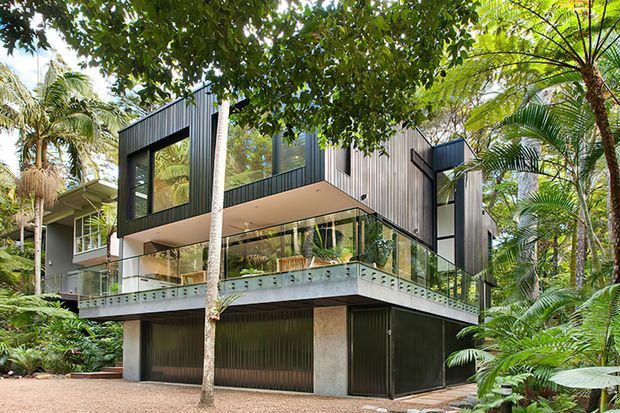Exterior view of a contemporary multi-level residence set within dense tropical landscaping. The house features dark vertical cladding, extensive glass walls and balustrades, projecting upper volumes, and a lower-level garage or enclosed storage area.