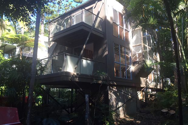 A multi-storey contemporary house elevated above a lush tropical garden, with dark cladding, extensive glazing, projecting balconies, and screened lower-level supports. The building is surrounded by dense vegetation and tall palms.