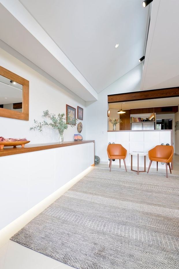 Bright open-plan interior featuring a white built-in sideboard with a timber benchtop, a large grey patterned rug, two orange leather lounge chairs and a small round table. A timber-framed mirror, artwork and decorative objects line the wall, while the background contains white cabinetry, open shelving, timber wall panelling and integrated lighting. The ceiling is high and angled, with recessed and track-style spotlights.