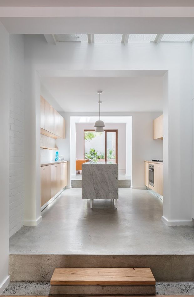Bright contemporary kitchen viewed through a wide opening, with pale timber cabinetry on both sides, a central stone island, polished concrete flooring, white-painted masonry walls, exposed roof framing and skylight-like overhead glazing. A rear window and indoor planting bring natural light into the space, while a short concrete step separates the foreground from the kitchen.