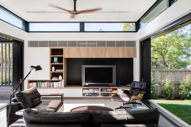 Bright contemporary living room with a large black leather sectional sofa, mid-century-style lounge chair, television and built-in timber entertainment cabinetry. Clerestory windows, extensive black-framed glazing, a ceiling fan and a garden outlook create an airy, light-filled space.