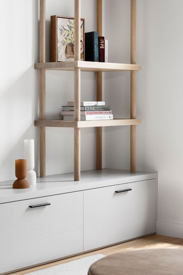 A bright, minimalist interior vignette featuring a low white built-in sideboard with simple black handles and an open timber shelving unit above it. The shelves display books and framed artwork, with small sculptural objects on the cabinet. Soft natural light, pale walls, light timber flooring and restrained neutral styling create a calm contemporary atmosphere.