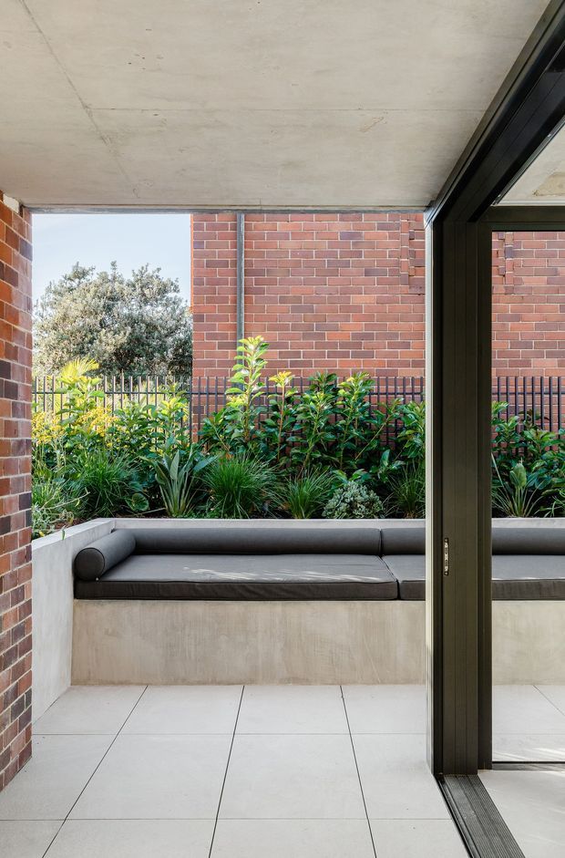 A contemporary covered courtyard or patio viewed from inside through a large dark-framed sliding glass door. The space features a built-in concrete bench with a grey upholstered cushion, pale large-format tiled flooring, exposed red brick walls, a raw concrete soffit, and a lush planted garden bed enclosed by a dark metal fence. The composition emphasises the connection between the interior and landscaped outdoor space.
