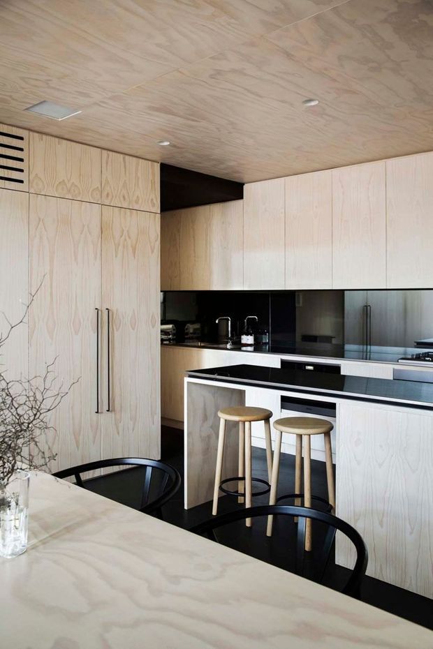 A contemporary kitchen dominated by pale plywood cabinetry and matching plywood ceiling and dining table. Full-height storage lines the left wall, while a central island with a dark benchtop provides seating for two on timber stools. Black reflective splashback panels, dark flooring, slim black handles, integrated appliances, and black dining chairs create strong contrast. The space has clean lines, recessed downlights, and a refined, minimalist material palette.