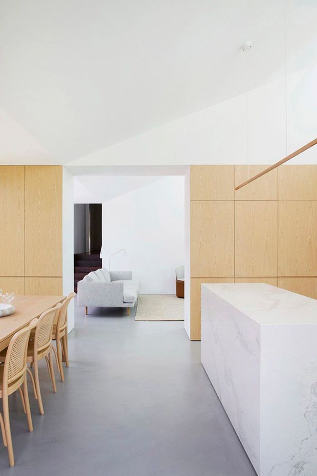 Bright minimalist open-plan kitchen and dining area with pale timber built-in cabinetry, a large white stone island, smooth grey flooring, and a light upholstered sofa visible beyond. A dining table with woven timber chairs occupies the foreground, while a simple timber stair handrail adds a linear architectural detail.