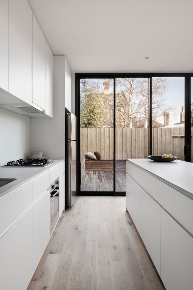 A bright, minimalist galley kitchen with white flat-panel cabinetry, integrated appliances, a gas cooktop, pale timber flooring, and large black-framed glazed doors opening to a timber deck and fenced outdoor area. A white island with a simple fruit bowl occupies the right side of the room.