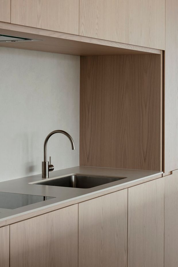 Close-up view of a minimalist kitchen featuring pale natural-wood cabinetry, a continuous wood overhead unit and side panels, a dark grey stone benchtop with an integrated stainless-steel sink, and a slim dark metal gooseneck tap. The backsplash is a smooth light neutral surface, with clean handleless joinery and restrained detailing.