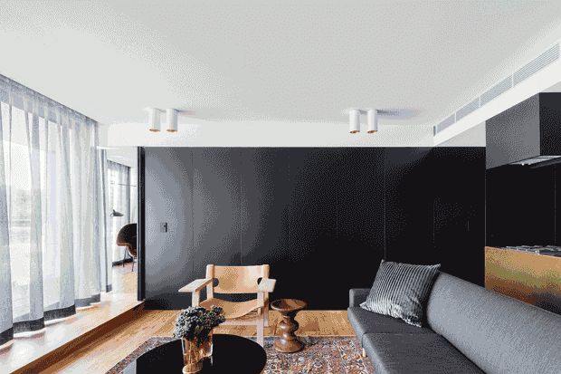 A refined contemporary living room with a dark feature wall, grey sectional sofa, sculptural timber lounge chair, round black coffee table, patterned rug, and timber flooring. Sheer floor-to-ceiling curtains line the glazed wall, bringing in abundant natural light. A partially visible open kitchen with dark cabinetry and a warm timber-toned island appears at right.