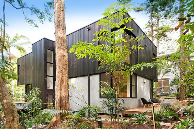 Contemporary two-storey house set among mature tropical landscaping, with a dark vertical metal-clad upper level, white rendered lower walls, large black-framed windows, and a steep gabled roof form.