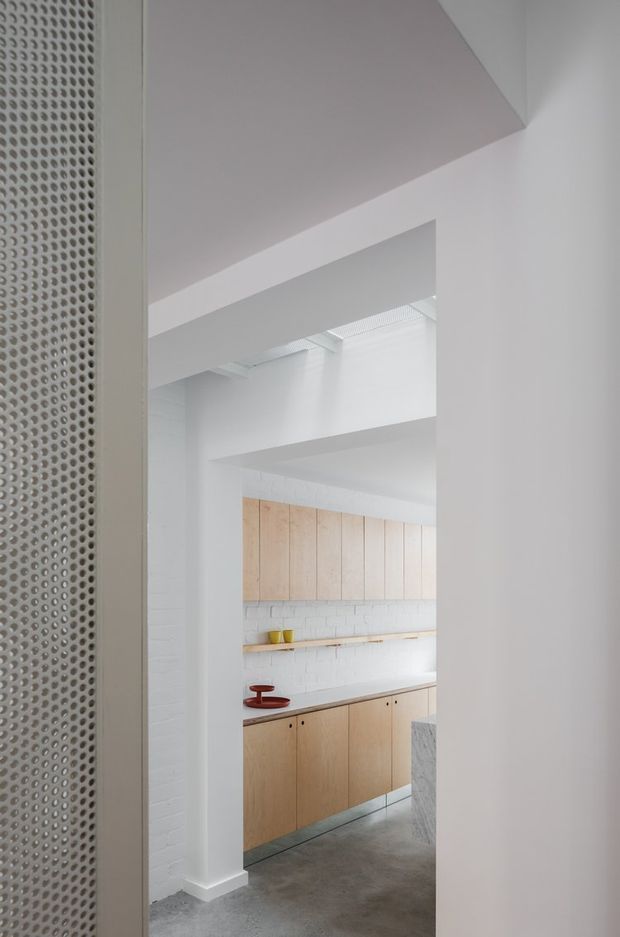 A minimalist interior view looking through a doorway into a bright kitchen. The kitchen features pale timber upper and lower cabinetry, a dark benchtop, white-painted brick walls, exposed ceiling structure, and a polished grey concrete floor. A perforated metal screen frames the left side of the composition.