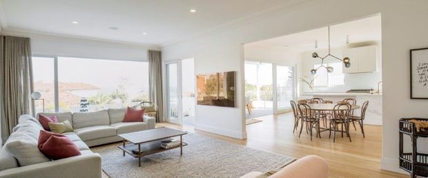 Bright, open-plan living and dining area with large corner glazing, a light neutral sectional sofa, timber flooring, a low coffee table, and a sculptural dining pendant. The dining zone opens toward a contemporary white kitchen, creating an airy and refined residential interior.