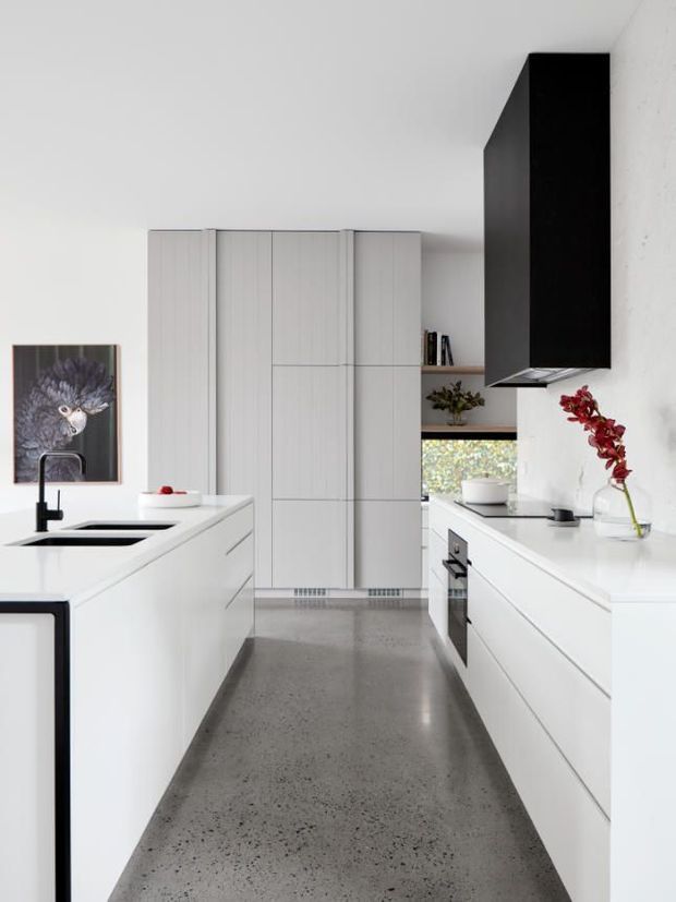 A bright, minimalist galley kitchen with a central white island and integrated sink, handleless white cabinetry, white benchtops, a contrasting black rangehood, and a polished grey concrete or terrazzo-like floor. A small dining or living area is glimpsed beyond the kitchen, with restrained decorative accents and abundant natural light.
