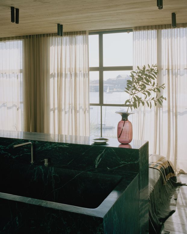 A refined open-plan interior with a deep green stone kitchen island in the foreground, featuring an integrated sink and waterfall-style sides. Floor-to-ceiling sheer curtains frame large dark-framed windows overlooking water and distant buildings. A pink glass vase with foliage, a sculptural bowl, and a draped throw add restrained decorative detail. The ceiling is clad in pale timber boards with cylindrical black pendant or track fixtures, and the overall atmosphere is elegant, warm, and softly lit.
