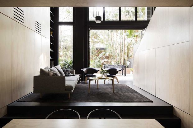 A refined contemporary living area with pale timber built-in wall panelling and cabinetry, a dark raised platform, large black-framed windows, and a compact seating arrangement around a coffee table. The room opens visually to a leafy outdoor courtyard or garden, with a dining table and chairs in the foreground. The palette is light timber, white, charcoal, and black, with strong natural light and clean geometric lines.