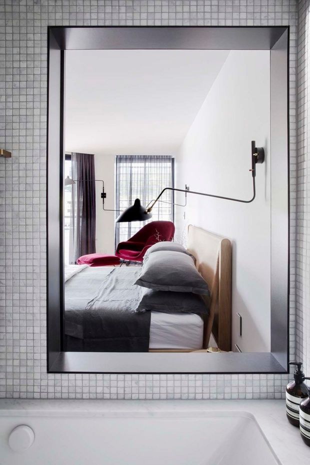A contemporary bathroom view dominated by a large black-framed rectangular mirror set into a wall of small square grey mosaic tiles. The mirror reflects an adjacent bedroom with a neatly made bed, pale timber headboard, sculptural red lounge chair, grey bedding, and a distinctive black articulated wall lamp. A white built-in bathtub occupies the foreground, with monochrome accessories at the right edge.