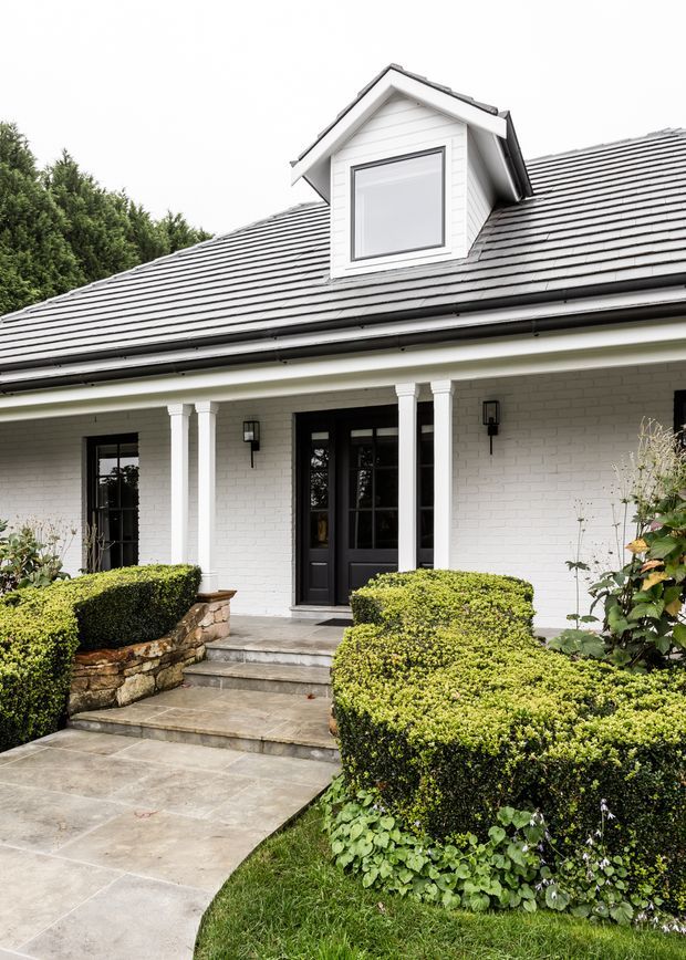 Front exterior view of a refined white-painted brick home with a dark tiled pitched roof and central dormer window. A covered entry porch is supported by paired white columns and reached by broad stone steps, with black-framed glazed doors, traditional wall lights, and carefully clipped hedges and stone garden edging framing the approach.