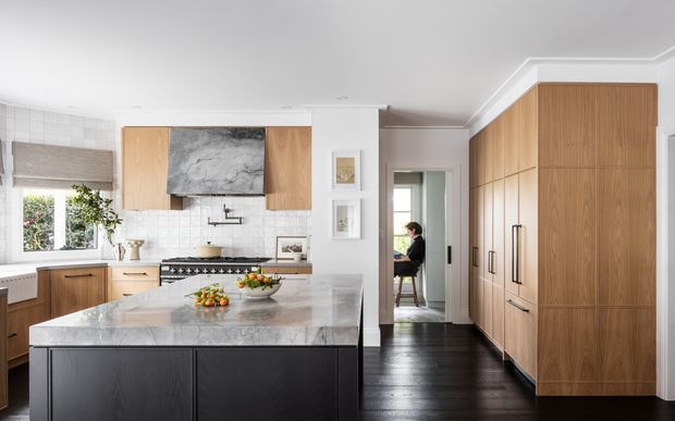 A refined contemporary kitchen with a large marble-look island, dark cabinetry, warm wood wall cabinetry, white tiled splashback, and black stained timber flooring. The room includes a freestanding range cooker, integrated appliances, extensive storage, and a small dining or sitting area visible through the doorway.