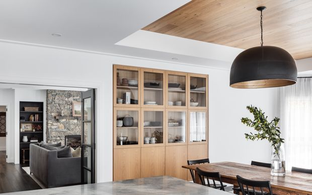 Bright contemporary open-plan dining and living area with a large built-in timber display cabinet, a substantial timber dining table, and a dramatic black dome pendant. A timber-lined ceiling recess adds warmth above the dining zone, while black-framed glazing opens to a living room with a stone feature wall and fireplace.