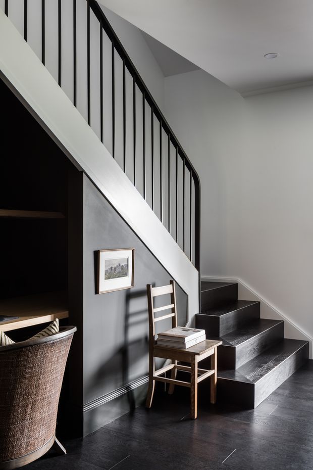 Interior view of a contemporary stairway with dark timber or charcoal-finished treads, slim black vertical balustrades, and a white stringer. The staircase is set against grey walls with dark flooring, with a small timber chair and side table beneath the stairs and built-in dark storage visible to the left.