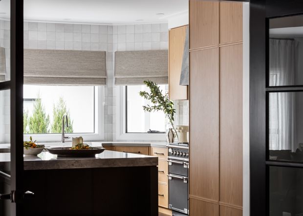 A bright contemporary kitchen viewed through a black-framed doorway. The room features a dark kitchen island with a stone benchtop, warm timber cabinetry extending to the ceiling, a freestanding stainless-steel range cooker, pale tiled walls, large windows with woven Roman blinds, and a small dining or utility area glimpsed beyond the black-framed glazed door.