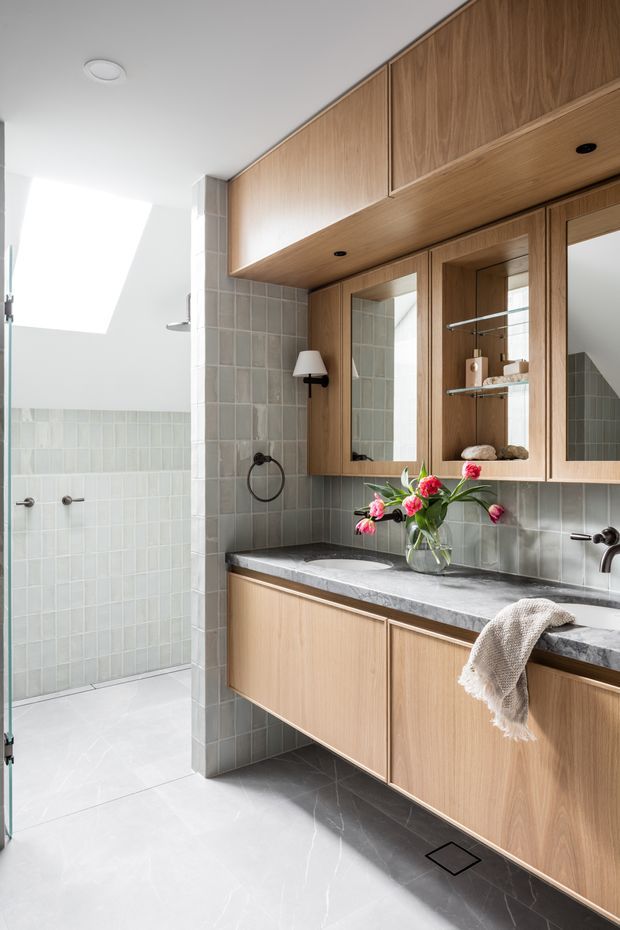 Bright contemporary bathroom featuring a floating light-wood vanity with integrated cabinetry, mirrored wall cupboards, a pale stone benchtop, grey-green tiled walls, and a walk-in shower separated by a tiled partition. Black tapware and hardware provide contrast, while a small vase of flowers adds colour.