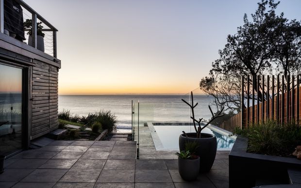 A modern coastal residence overlooking the ocean at sunset, with dark timber-clad exterior walls, expansive glazing, a paved terrace, glass balustrades, built-in planter beds, and a swimming pool aligned with the sea view.