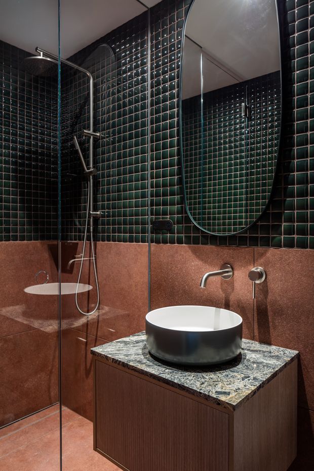 A compact, moody contemporary bathroom with a walk-in glass shower, dark glossy green square wall tiles, and terracotta-toned wall and floor tiles. A round black vessel basin sits on a dark timber vanity with a richly veined stone benchtop. Brushed metal wall-mounted tapware, a large rounded mirror, and integrated shower fixtures complete the refined, dramatic interior.