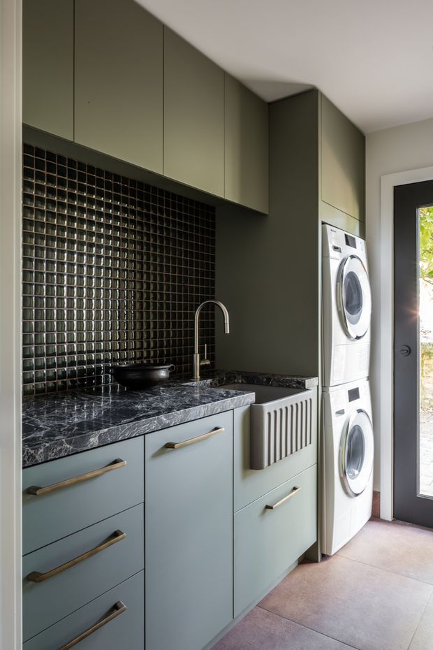 Compact, highly considered laundry room with full-height muted green cabinetry, dark marble-look benchtop, glossy black square tile splashback, integrated sink and brass-toned handles. A stacked white washer and dryer sit beside a dark-framed glazed exterior door, while a heated towel rail is mounted to the cabinetry. The room has a refined, moody and contemporary feel.