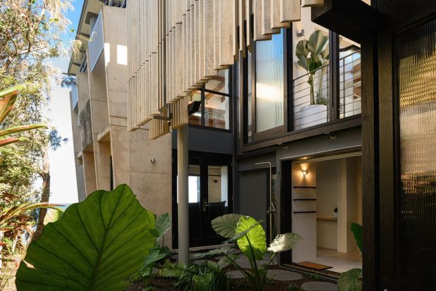 Contemporary multi-level residential facade viewed from a landscaped courtyard, featuring dark framed glazing, textured vertical concrete fins, projecting window hoods, and recessed entry areas. Tropical foliage softens the architectural composition, while warm interior lighting is visible through the openings.