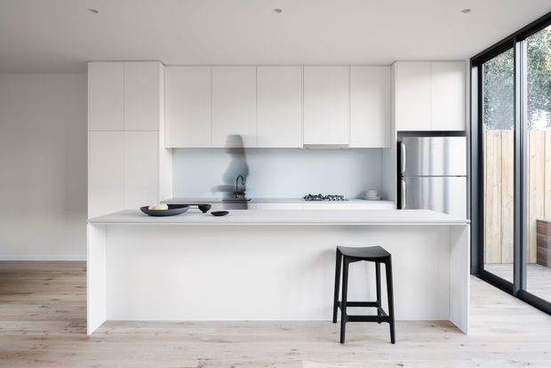 Bright, minimalist open-plan kitchen with a large white island, integrated white cabinetry, pale backsplash, stainless-steel refrigerator, gas cooktop, and a single black bar stool. Full-height black-framed glazing opens toward an exterior deck or courtyard, while pale timber flooring adds warmth.