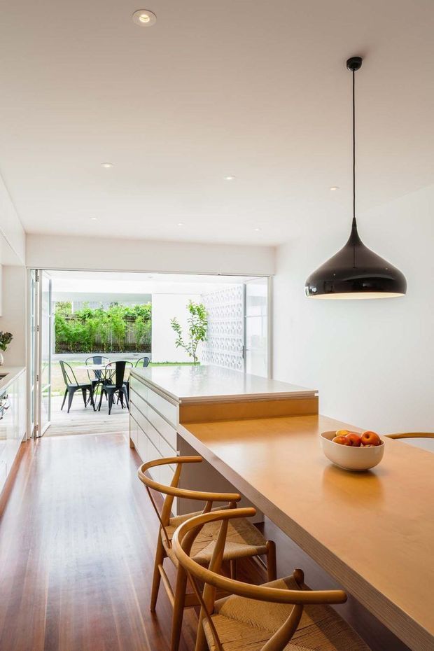 Bright contemporary kitchen viewed from the island toward a large opening to an outdoor dining area. The room features warm timber flooring, a large light timber island with integrated white cabinetry, a black sculptural pendant light, white walls and ceiling, and extensive sliding glazing. Wooden dining stools line the island, while a patterned privacy screen and greenery are visible outside.