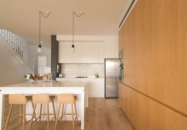 Bright contemporary kitchen with a large white island and three timber bar stools, white flat-panel cabinetry, a white subway-tile splashback, stainless-steel appliances, and extensive warm timber built-in cabinetry along the right wall. A staircase and upper-level railing are visible to the left, with simple pendant lights and light timber flooring creating an open, airy feel.