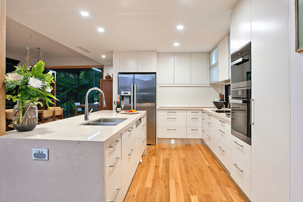 Bright contemporary kitchen with a large white engineered-stone island, integrated sink and curved chrome tap, extensive white handleless and flat-panel cabinetry, stainless-steel appliances, and warm timber flooring. Full-height glazing and an adjoining dining/living area are visible beyond the island, with recessed ceiling lighting and a floral arrangement adding contrast.