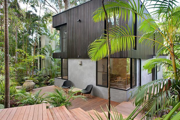A contemporary two-storey dwelling nestled in dense tropical landscaping. The building combines dark vertical cladding on the upper level with a smooth light-grey rendered lower level, large black-framed windows, and a timber deck and walkway winding through lush palms and planted garden beds. Outdoor lounge chairs sit beside the deck.