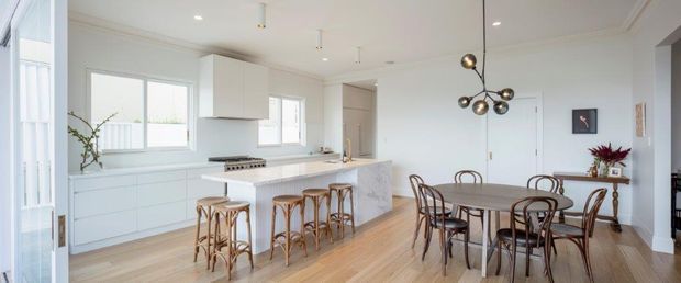 Bright open-plan kitchen and dining area with white built-in cabinetry, a pale stone island, timber flooring, and a round dining table with bentwood chairs. Large windows provide abundant natural light, while a sculptural multi-globe pendant adds a dark metallic accent.