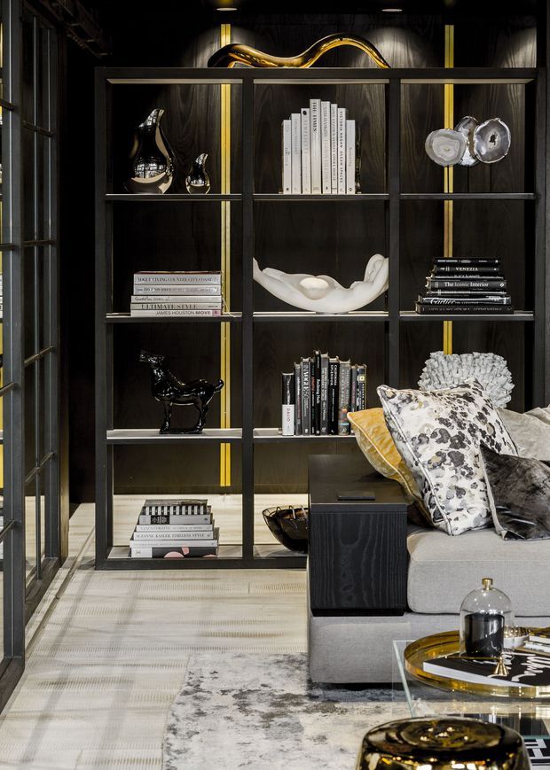 A sophisticated, dark-toned living room featuring a full-height black metal and dark timber display shelving unit with integrated warm yellow vertical accents. The shelving holds books, sculptural objects and decorative ceramics. A grey upholstered sofa with patterned cushions sits in the foreground alongside glass and dark metal coffee tables. The room has layered neutral textures, patterned rugs, dramatic low lighting and a refined, curated aesthetic.