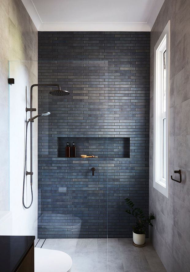 A contemporary walk-in shower bathroom with a dark charcoal brick-look tiled feature wall, recessed shower niche, black rain shower and handheld fittings, pale grey wall and floor tiles, and a narrow white-framed window. A partial white freestanding bath and dark vanity are visible in the foreground, with a small potted plant adding greenery.