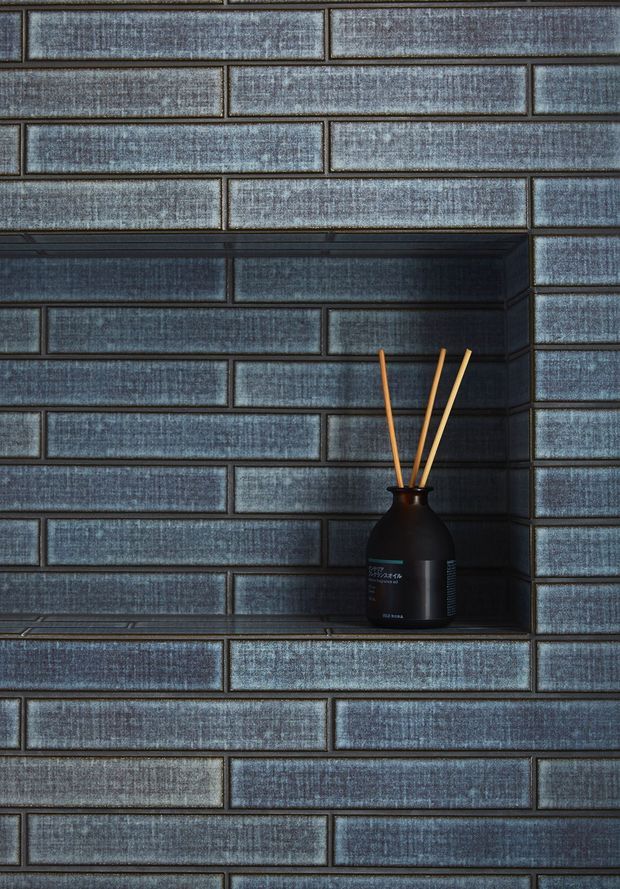 Close-up of a dark blue-grey textured rectangular tile wall in a running-bond brick pattern, featuring a recessed tiled niche with a black diffuser bottle and wooden reeds. The image focuses on the tile finish and built-in wall recess, with a moody, refined appearance.