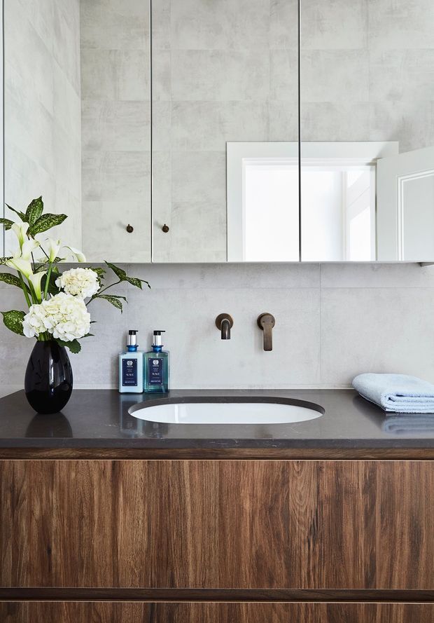 A refined contemporary bathroom vanity with a floating timber-look cabinet, dark stone benchtop, integrated white oval basin, wall-mounted bronze tapware, and large mirrored cabinetry. Pale grey concrete-look wall tiles create a clean, modern backdrop, complemented by folded towels and a floral arrangement.