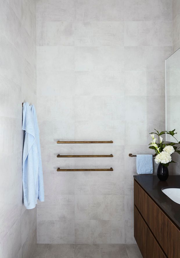A contemporary bathroom with large pale grey concrete-look wall and floor tiles, three horizontal bronze towel rails, a wall-mounted light timber vanity with dark benchtop, a white basin, and a large mirror. Pale blue towels and a vase of flowers add soft colour.