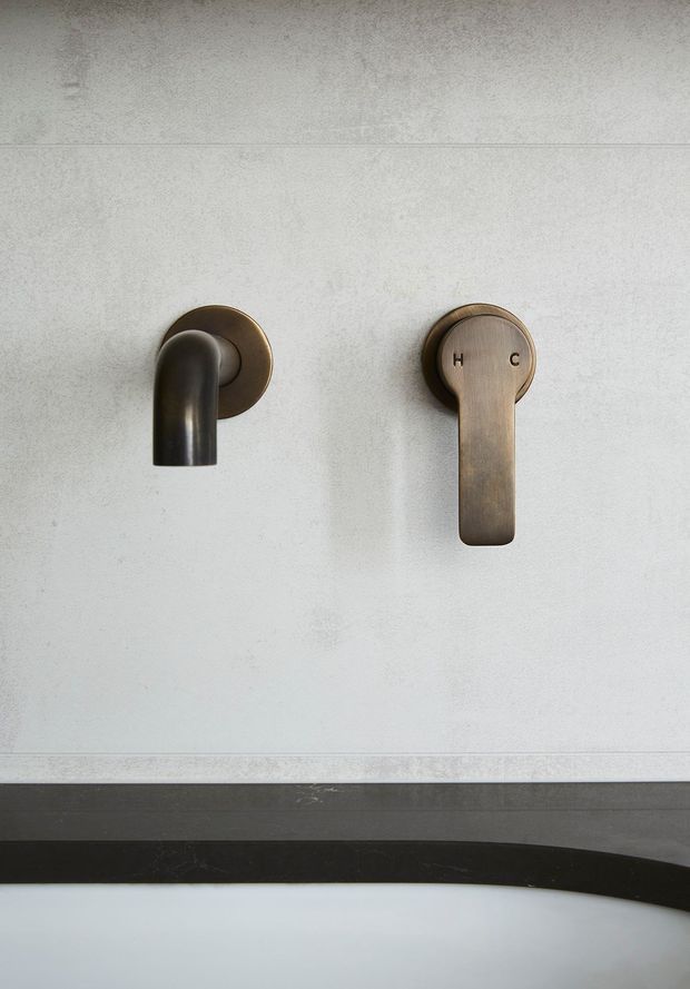 Close-up of a minimalist wall-mounted bathroom tap set, featuring a dark curved spout and a separate rounded lever control in a warm bronze or brushed metal finish. The fittings are installed on a pale textured tiled wall above the edge of a dark bathtub or basin.