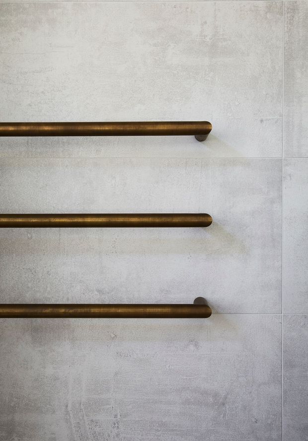 Close-up detail of three horizontal bronze-toned metal rails mounted against a pale grey, textured tiled wall. The minimalist composition emphasizes the contrast between the warm metal hardware and cool concrete-like wall finish.