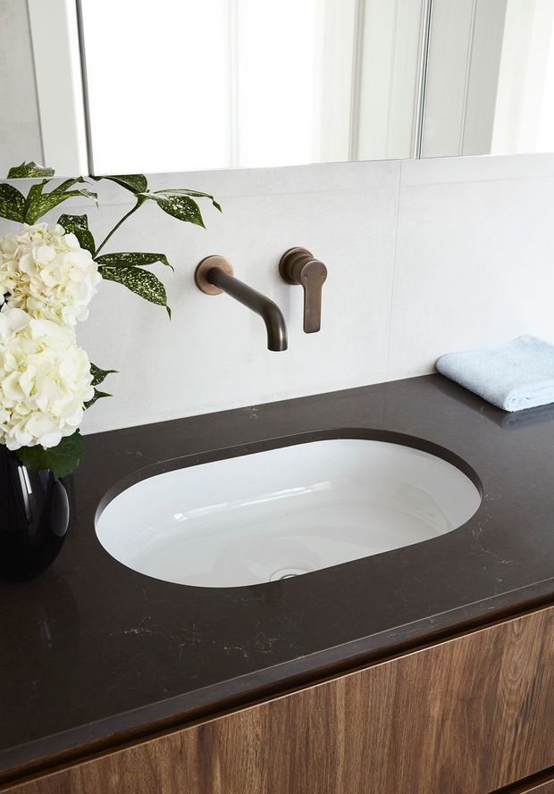 Close-up of a refined bathroom vanity featuring a white inset oval basin set into a dark stone benchtop, timber-grain vanity cabinetry, a bronze wall-mounted tap set, pale tiled wall, and a bright window above. A vase of white flowers and folded towel provide limited styling accents.