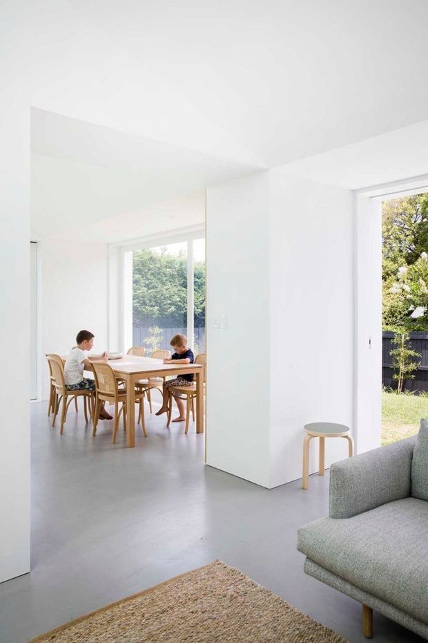 Bright minimalist open-plan interior viewed from a living room toward a dining area. White walls, broad openings, large glazed doors and windows, a smooth grey floor, pale timber dining furniture, and a textured neutral rug create a clean, airy space. A grey upholstered sofa and small round stool are visible in the foreground, while two children sit at the dining table. The garden is visible through the glazing.