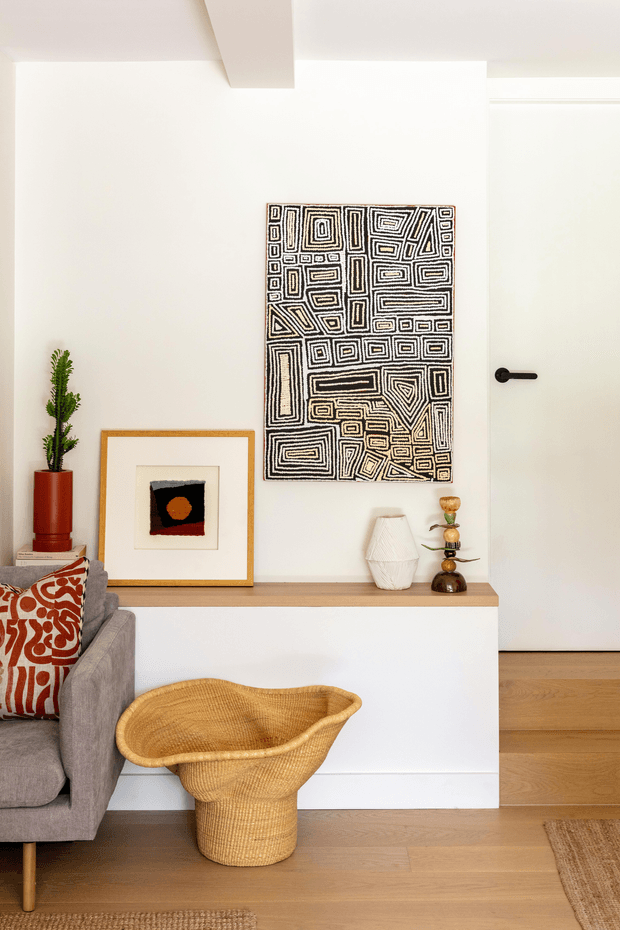 A bright, calm contemporary living room vignette with white walls, warm timber flooring, and a minimalist built-in white console. The scene includes a grey upholstered sofa with a bold rust-and-cream patterned cushion, a woven rattan basket, a tall potted cactus, framed artwork, sculptural ceramics, and a white door with a black lever handle. A geometric black-and-white wall artwork provides a graphic focal point.