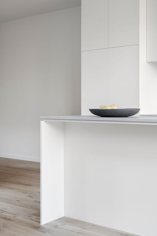 A minimalist kitchen interior focused on a white island or peninsula with a pale grey stone-look benchtop and integrated white cabinetry. The space has plain white walls, light timber flooring, clean slab-front joinery, and a dark shallow bowl with fruit as the only visible decor. The composition is bright, sparse, and highly contemporary.