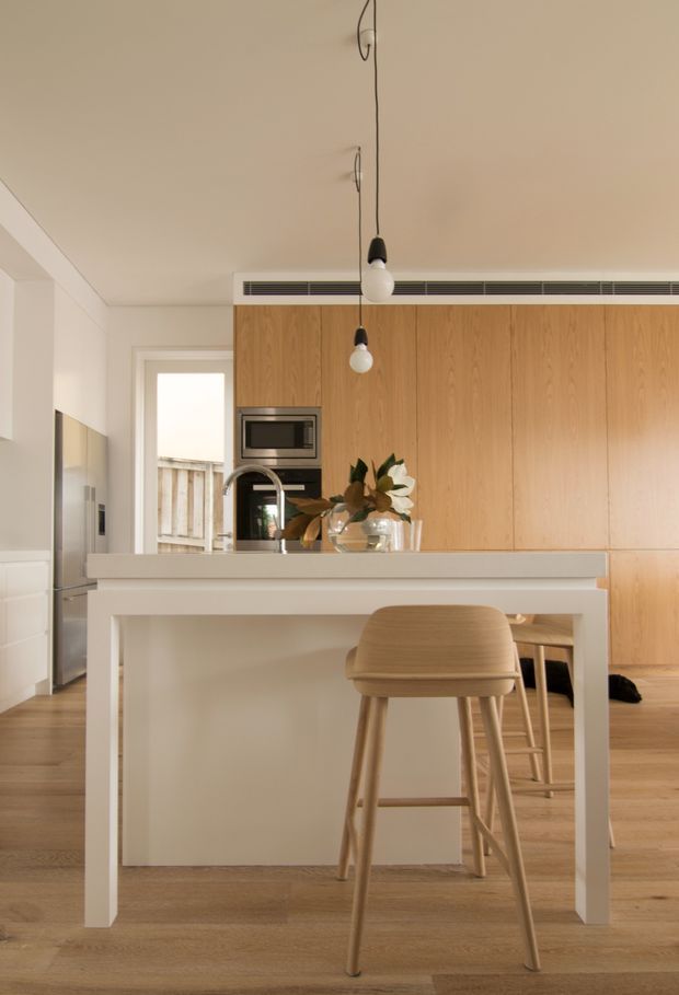 Warm, minimalist kitchen interior viewed from the dining side, featuring a large white island with integrated seating, pale timber flooring, full-height timber cabinetry, built-in stainless appliances, and a linear concealed air-conditioning vent. Two simple pendant lights hang above the island, with natural light entering through a rear doorway and window.