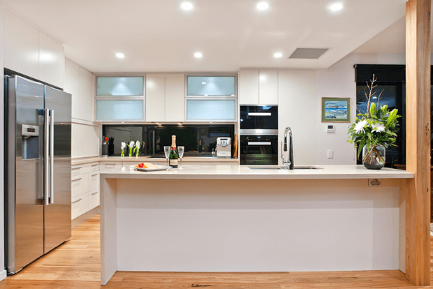 Bright contemporary kitchen with a large white island, integrated stainless-steel appliances, white flat-panel cabinetry, glossy black splashback, timber flooring, and a timber structural post at the right. A vase of flowers and small countertop items provide limited decorative accents.