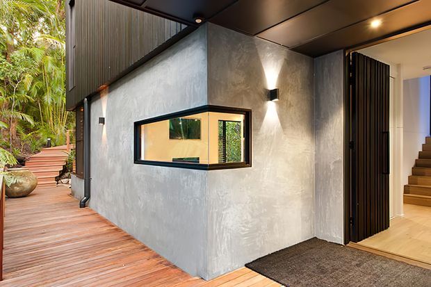 Exterior approach to a contemporary residence, featuring a textured grey rendered wall, black-framed horizontal window, dark metal soffit and screening, and a timber deck walkway leading through lush tropical landscaping. An open entry reveals a warm timber staircase and light interior flooring.