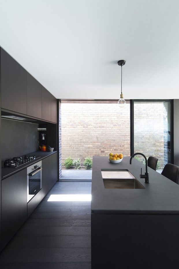 A compact contemporary kitchen with a dark, streamlined galley layout and large central island. Matte charcoal cabinetry and integrated appliances line the left wall, while the island incorporates a deep stainless-steel sink with a tall black tap and seating for several people. A full-height glazed opening at the rear overlooks a small brick courtyard, bringing strong natural light into the otherwise moody interior. A single pendant hangs above the island, and dark timber flooring runs throughout.